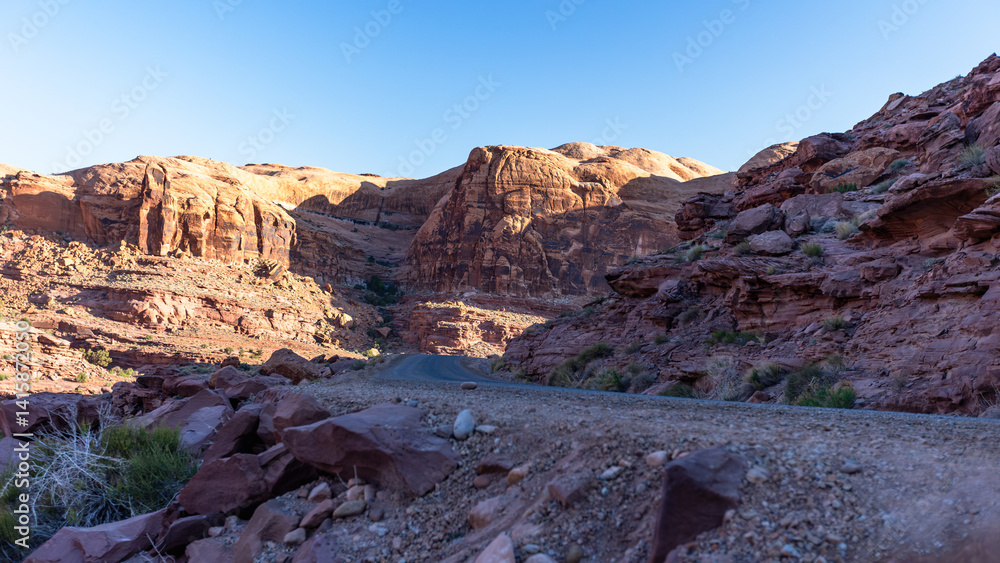 Fototapeta premium Early morning drive through the rugged dirt roads of Moab, Utah, with golden sunlight casting dramatic shadows across red rock canyons and remote desert landscapes