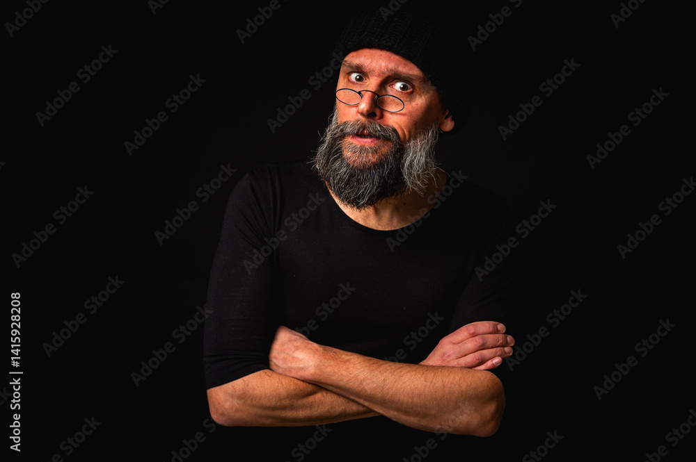 Stylish portrait of a confident mature man with his arms crossed in front of him, wearing a knitted hat and glasses. Staring, gray beard and black vest. Stylish discreet portrait on a dark background,