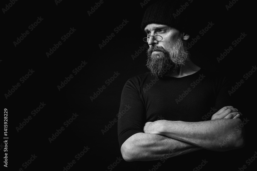 Black and white, stylish portrait of a confident mature man with his arms crossed in front of him, wearing a knitted hat and glasses. Staring, gray beard and black vest. Stylish discreet portrait