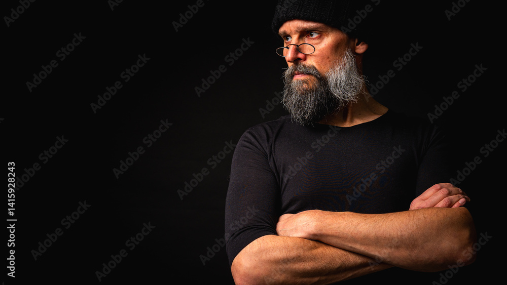 Stylish portrait of a confident mature man with his arms crossed in front of him, wearing a knitted hat and glasses. Staring, gray beard and black vest. Stylish discreet portrait on a dark background,