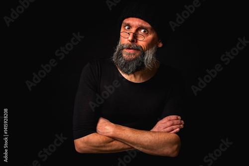 Obraz na płótnie Stylish portrait of a confident mature man with his arms crossed in front of him, wearing a knitted hat and glasses. Staring, gray beard and black vest. Stylish discreet portrait on a dark background, w salonie