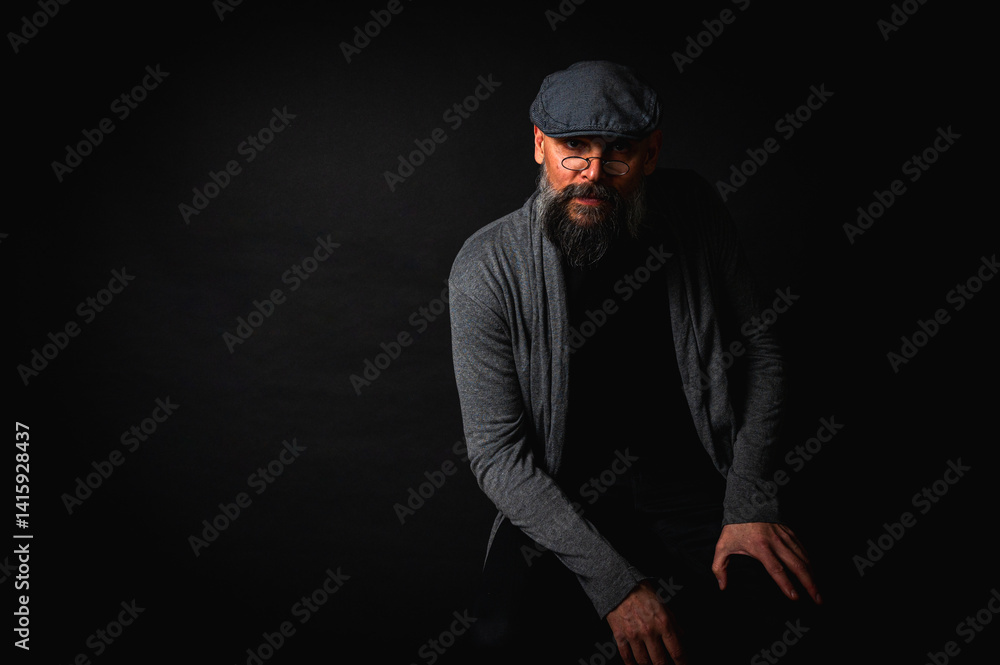 Close-up portrait of a serious bearded man in flat cap and glasses on black background. Stylish, intense look with confident and mysterious expression