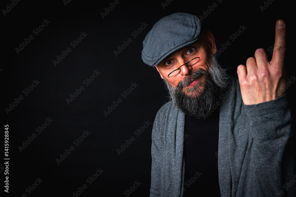 Elderly bearded man in cap and glasses shows a finger up gesture, expressing sarcasm or rebellion. Studio portrait on black background