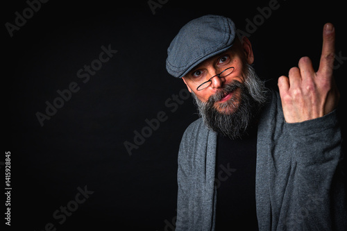 Obraz na płótnie Elderly bearded man in cap and glasses shows a finger up gesture, expressing sarcasm or rebellion. Studio portrait on black background w salonie