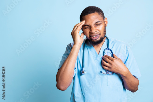 Fatigued tired nurse suffering from painful migraine after tiresome day at work, standing against blue background. Hospital staff affected by distressing headache during demanding night shift.