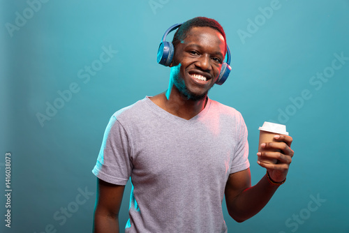 African american male individual stands with a disposable cup, listening to music on his wireless headset and looking at camera. Portrait of young man posing with coffee in his hand and headphones.