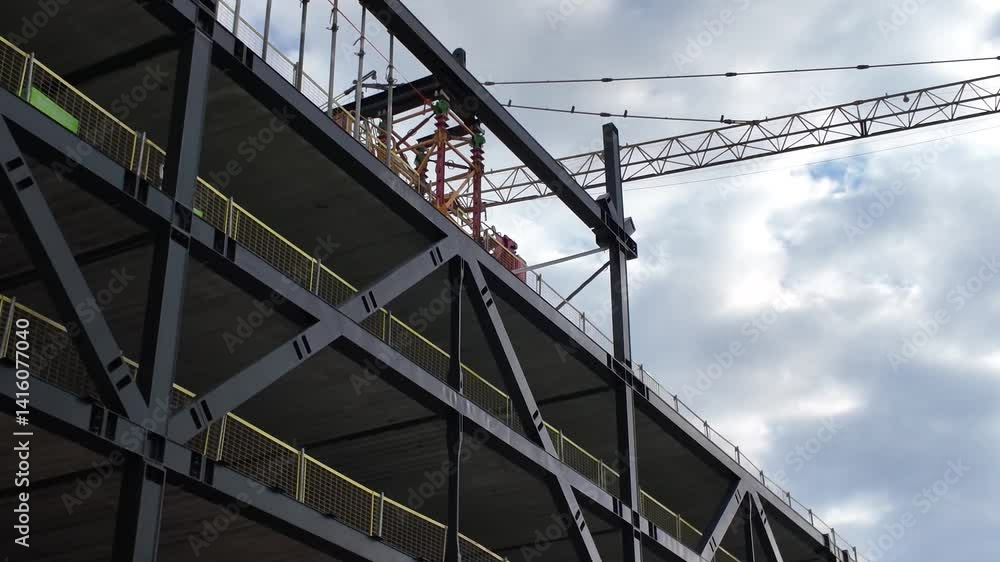 Partial view of a building under construction featuring exposed steel framework, safety railings, and a crane against a cloudy sky. Partial view of a building under construction featuring exposed steel framework, safety railings, and a crane against a cloudy sky.