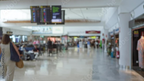 Blurred background of busy airport terminal with defocused travelers, departure boards, and shops creating a bokeh effect.