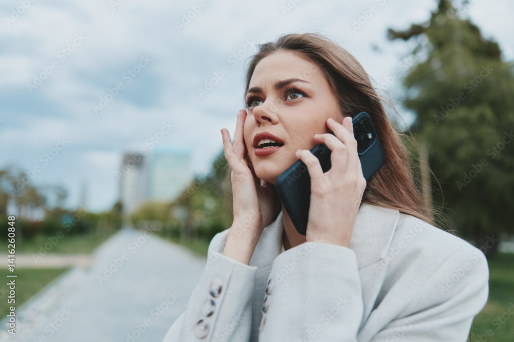Worried young woman in white blazer speaking on phone outdoors expressing stress and anxiety, urban background with green trees and cloudy sky Worried young woman in white blazer speaking on phone outdoors expressing stress and anxiety, urban background with green trees and cloudy sky