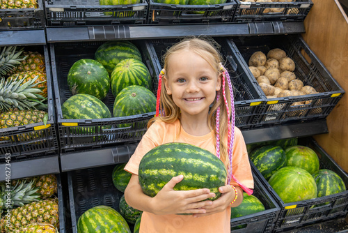 Cheerful little blonde girl holds a watermelon in fresh food supermarket. She stands in fruits and vegetables section with smile. Healthy eating. Family grocery shopping