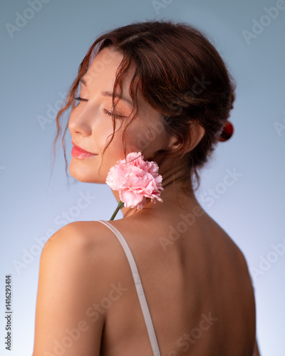 Obraz na płótnie Delicate studio portrait of a young woman with closed eyes, holding a gentle pink carnation on her shoulder. Her glowing skin and peaceful expression blend seamlessly with the pastel blue background w salonie