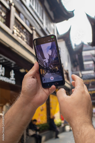 Man's hands holding smartphone, recording lively street view in traditional Chinese market. Blogger filming vivid red buildings and ornate roofs, perfect for a travel blog