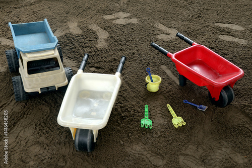 Toy wheelbarrows, a plastic dump truck, shovels, rakes, and a bucket on sand. Children's play scene outdoors or in an early stimulation space.