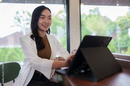 Free Download Smiling young asian businesswoman is using a tablet in a modern office with a window view of trees, working on a project and enjoying the natural light - GFXLoad