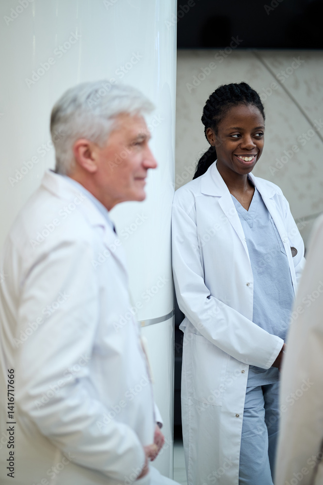 Medical professionals in conversation while standing in hospital. Smiling female doctor wearing white coat and scrubs. Male doctor with gray hair listening attentively Medical professionals in conversation while standing in hospital. Smiling female doctor wearing white coat and scrubs. Male doctor with gray hair listening attentively