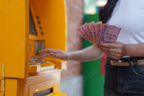 Obraz na plátně Woman withdrawing cash from an atm, holding thai baht banknotes in her hand, man