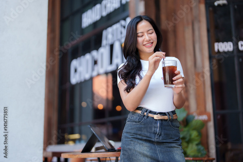 Fotografía Stylish young asian woman stands outside a cafe, savoring an iced coffee while s