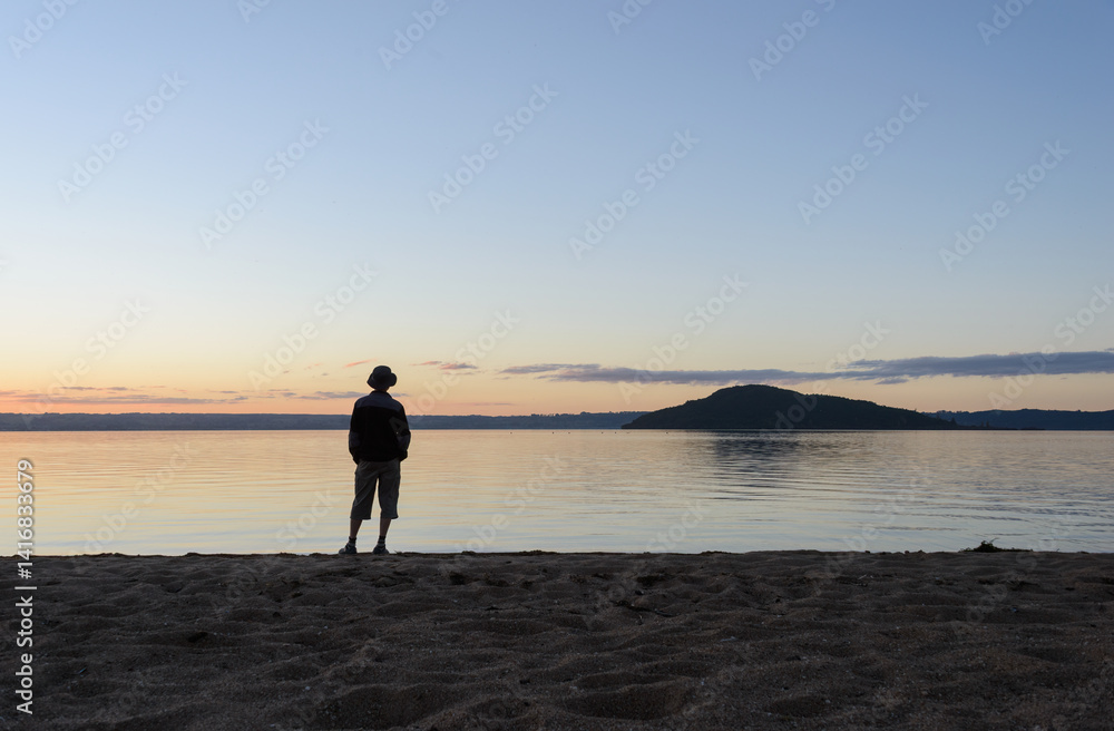 Man standing along on the beach at sunset. Mokoia Island in the distance. Lake Rotorua.