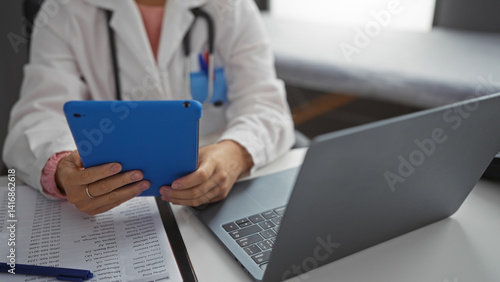 Obraz na płótnie Female doctor using tablet in clinic room with stethoscope nearby, managing records on laptop at workplace emphasizing healthcare and technology integration. w salonie