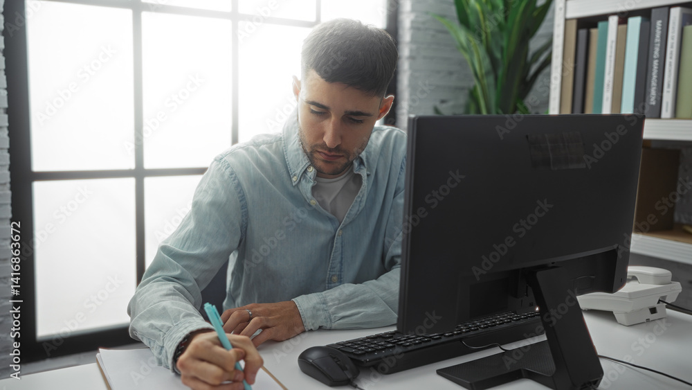 Man working indoors in office with computer and plant, focused on writing at a modern desk, daylight streaming through window onto bookshelves in background.