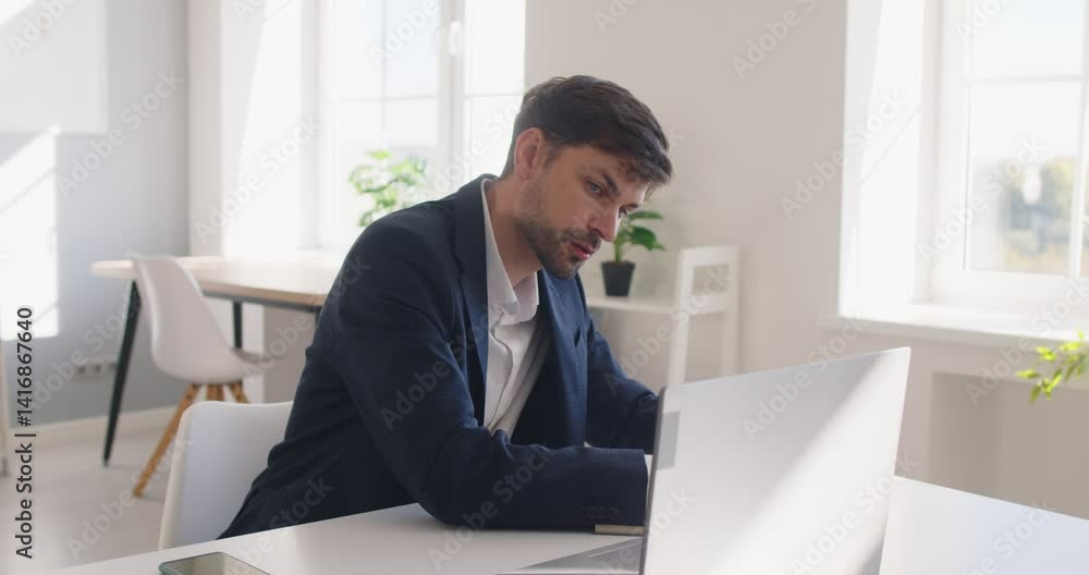 Focused business man sitting at desk on workplace, writing notes on work issues at notebook. Serious employee working on laptop, concentrating on tasks in bright and modern workplace environment. Focused business man sitting at desk on workplace, writing notes on work issues at notebook. Serious employee working on laptop, concentrating on tasks in bright and modern workplace environment.
