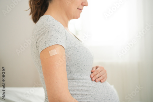 Image of a pregnant woman holding their upper arm with a bandage applied after receiving a vaccination. The scene emphasizes the importance of health care and protection.