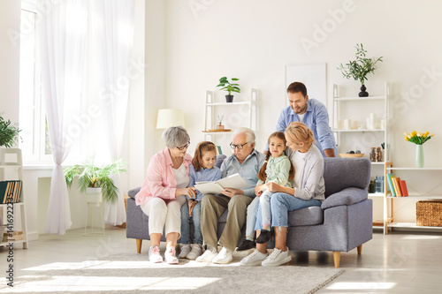 Multi generational extended family reading story together. Grandfather, grandmother, mom, dad and children sitting on sofa in light living room interior in grandparents' apartment and reading book