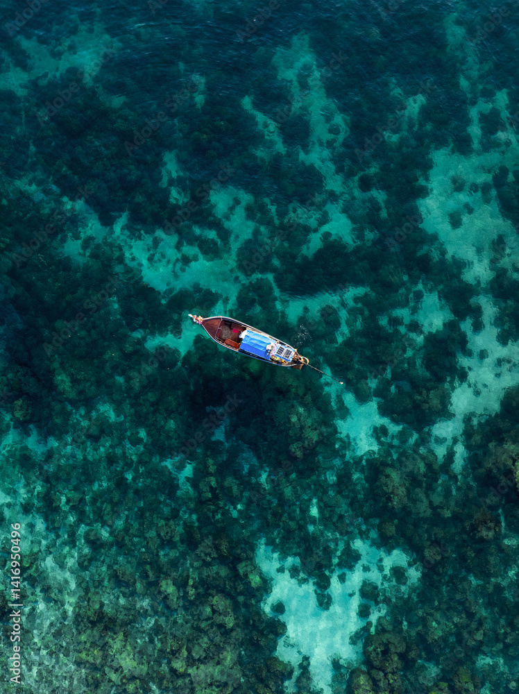 Wooden boat floating above a shallow coral reef near rocky formations in crystal-clear blue water in Thailand.