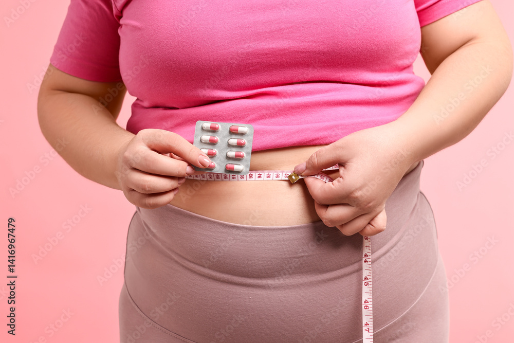 Plus size woman measuring waist with tape and holding blister of weight loss supplements on pink background, closeup