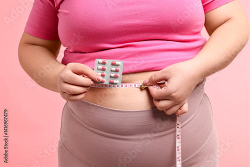 Obraz na płótnie Plus size woman measuring waist with tape and holding blister of weight loss supplements on pink background, closeup w salonie