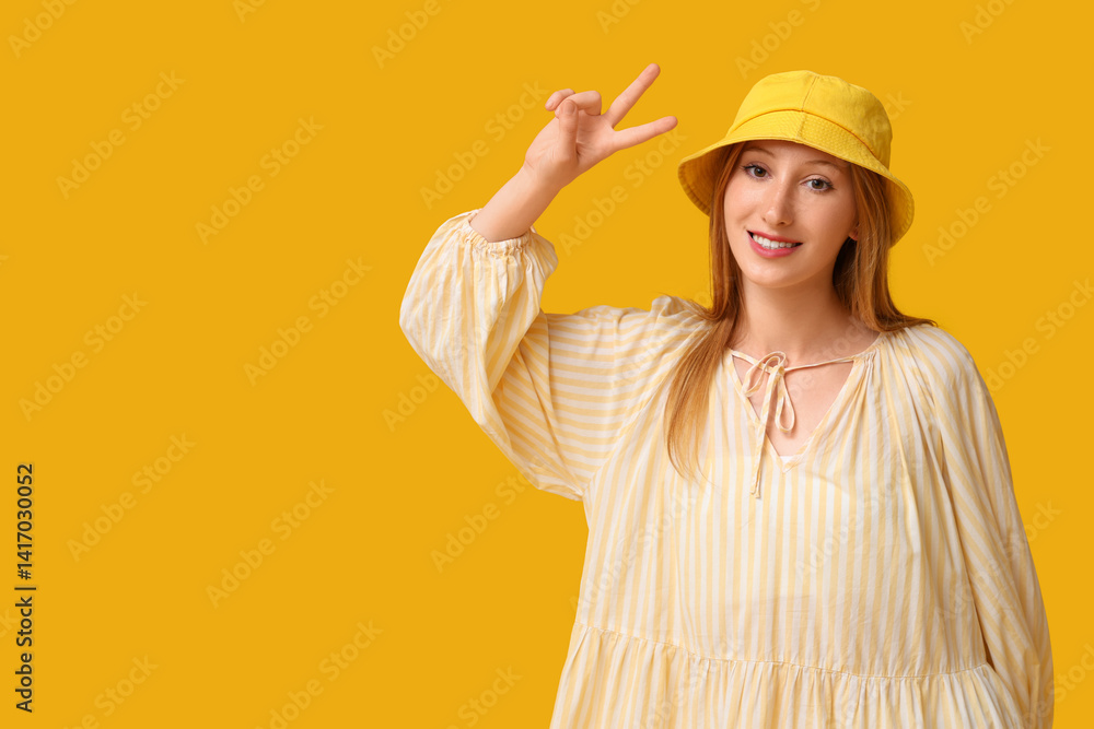 Beautiful young happy woman in stylish outfit with summer hat showing victory gesture on yellow background Beautiful young happy woman in stylish outfit with summer hat showing victory gesture on yellow background