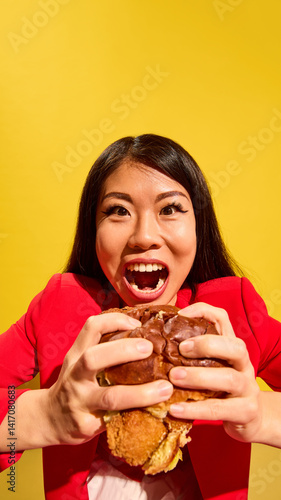 Obraz na płótnie Closeup of joyful Asian woman expressing excitement holding big burger against bright yellow background. Fast food restaurant poster. Concept of food, pop art, human emotions. Vertical layout w salonie