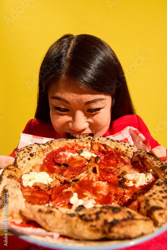 Woman in red bites giant pizza slice with intense hunger and playful face against bright yellow background. Delicious Italian cuisine. Concept of food, pop art, human emotions. Close-up