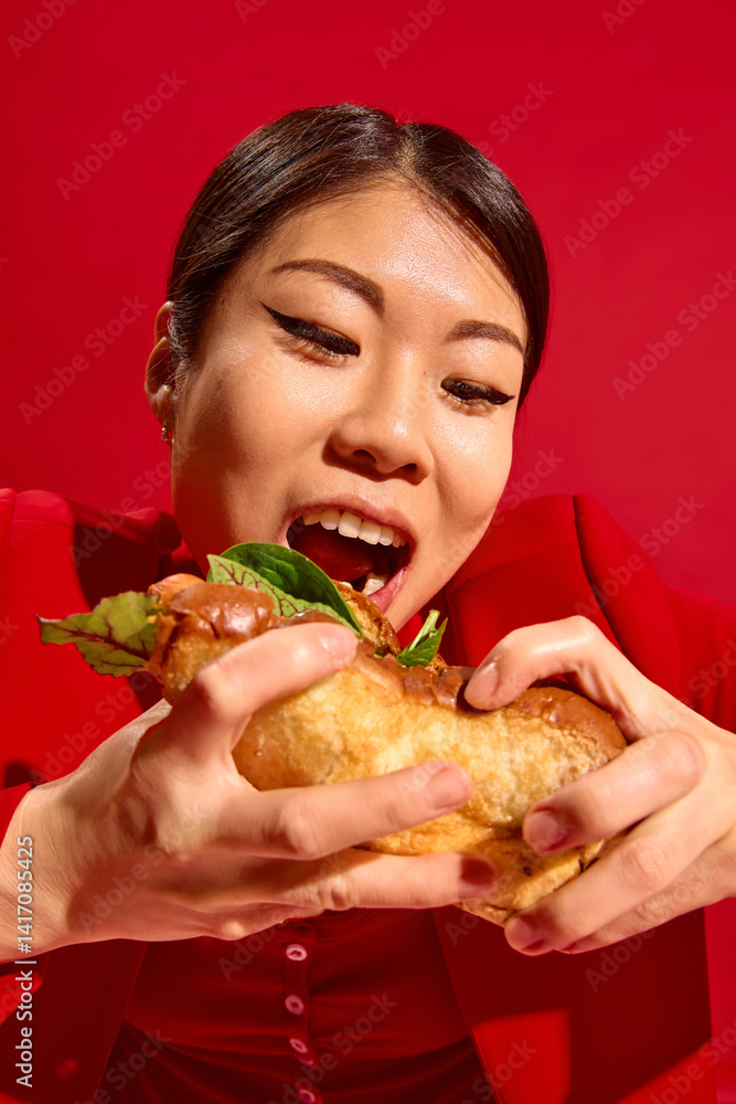 Delighted woman holds large sandwich with both hands and prepares for first bite, radiating playful emotion against red background. Concept of food culture, pop art photography, human emotions