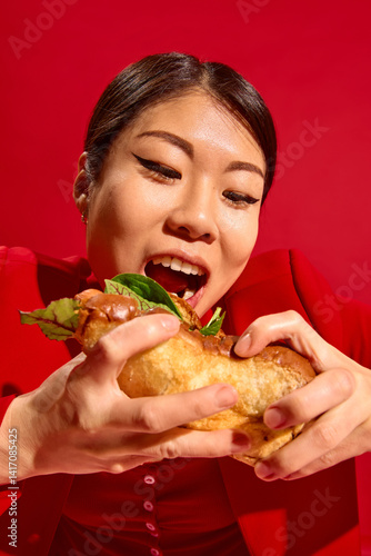 Delighted woman holds large sandwich with both hands and prepares for first bite, radiating playful emotion against red background. Concept of food culture, pop art photography, human emotions