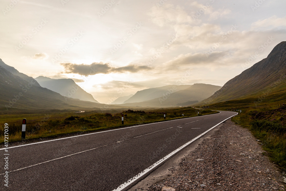 Naklejka premium Golden sunset over calm lakes and clear water reflections in The Trossachs and Loch Lomond. Long winding roads cross green hills and mountains in a peaceful, awe-inspiring natural landscape. Exploring