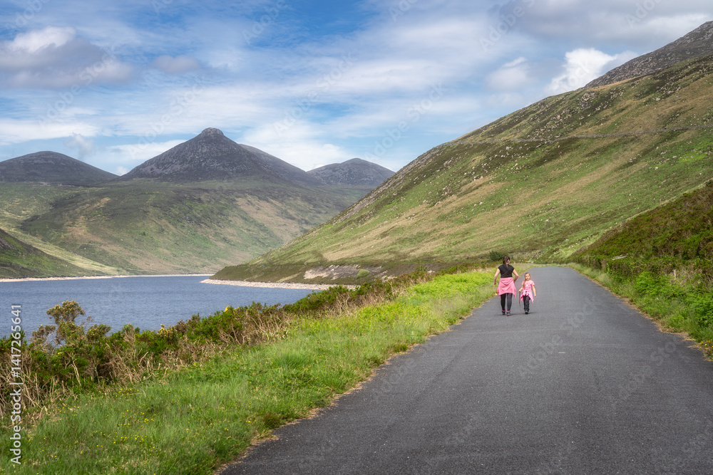 Mother and her joyful child leisurely stroll along Silent Valley Reservoir path that is beautifully surrounded by majestic Mourne Mountains and lush, vibrant greenery all around them, Northern Ireland
