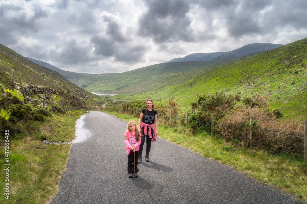 Mother and her daughter happily stroll along a picturesque pathway that is beautifully surrounded by vibrant greenery and lush rolling hills of Mourne Mountains beneath a cloudy sky, Northern Ireland