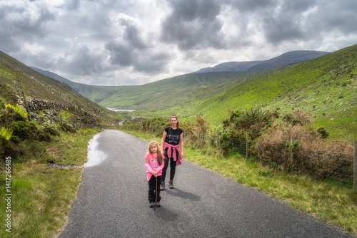 Mother and her daughter happily stroll along a picturesque pathway that is beautifully surrounded by vibrant greenery and lush rolling hills of Mourne Mountains beneath a cloudy sky, Northern Ireland