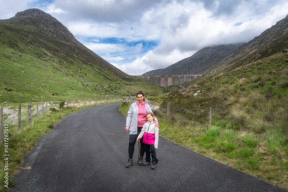 A joyful mother and her child happily pose together on a picturesque nature trail, surrounded by the lush greenery of Mourne Mountains and under a clear blue sky, Northern Ireland