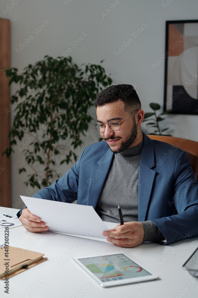Vertical portrait of smiling businessman reading documents at desk in modern office with sleek interior design Vertical portrait of smiling businessman reading documents at desk in modern office with sleek interior design