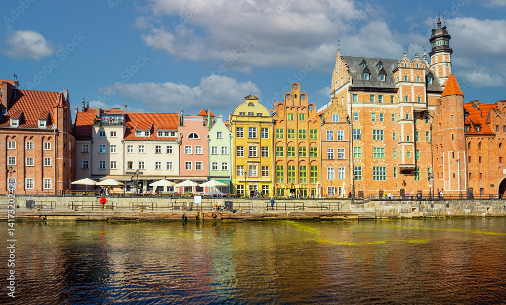Fototapeta premium Charming historic architecture lines the waterfront of Gdansk under a clear blue sky. The river is bustling with boats, offering a vibrant scene with the city's iconic brick buildings