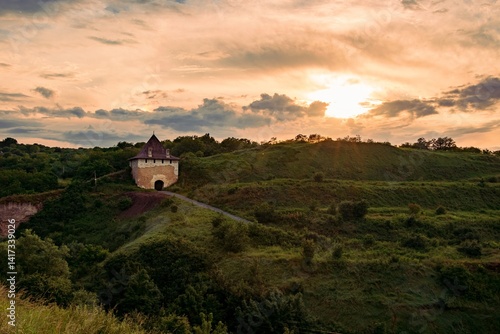 Watchtower of the architectural ensemble of the majestic medieval Khotyn fortress in Ukraine. Old stone tower with a pointed roof nestled on a grassy hill under a dramatic sky at sunset.