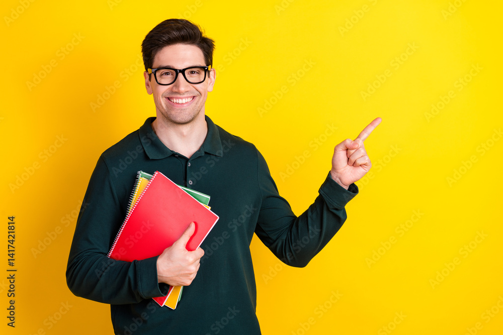 Young man in green polo holding notebooks while pointing to the side against a vibrant yellow background, smiling confidently