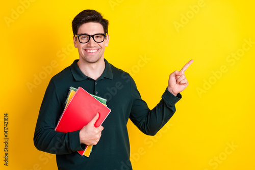Obraz na płótnie Young man in green polo holding notebooks while pointing to the side against a vibrant yellow background, smiling confidently w salonie