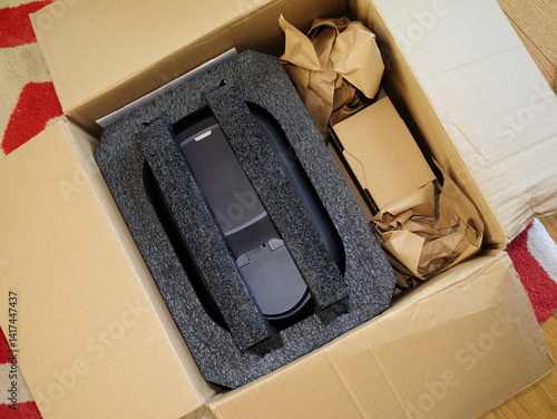 Opened cardboard shipping box containing a securely packed electronic device with foam padding and crumpled paper for protection placed on a patterned carpeted floor