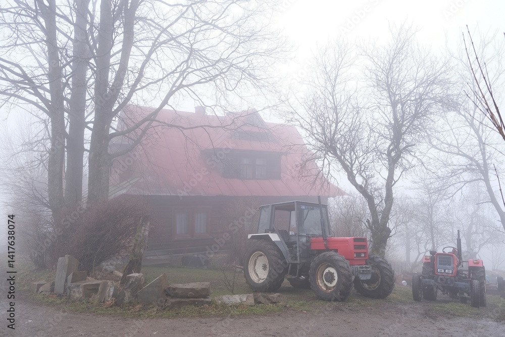 Very misty scenery tractor standing next to farm building in village.