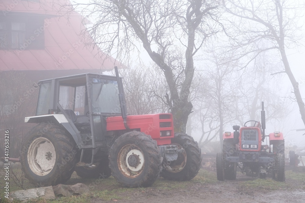 Very misty scenery tractor standing next to farm building in village.