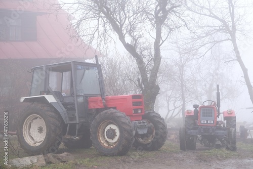 Very misty scenery tractor standing next to farm building in village.