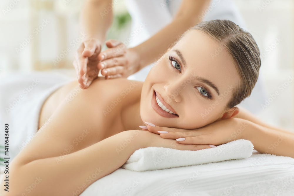 Portrait of a pretty happy smiling young woman relaxing in spa salon getting massage and looking cheerful at camera. Therapist doing back treatment procedure. Wellness and beauty day concept. Portrait of a pretty happy smiling young woman relaxing in spa salon getting massage and looking cheerful at camera. Therapist doing back treatment procedure. Wellness and beauty day concept.